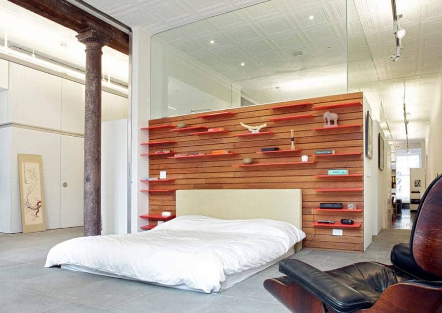 The white cushion is directly placed on the gray concrete flooring. This bed has a beige headboard that goes well with the wooden plank finish of the wall behind it with floating shelves on it for decors and to serve as bedside table.