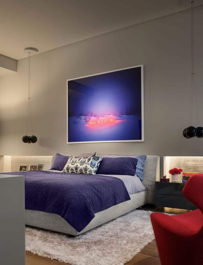 The light gray headboard of the bed is against a gray concrete wall that is adorned with an artwork that glows with neon lights. This is flanked by a couple of spherical pendant lights over modern bedside drawers.