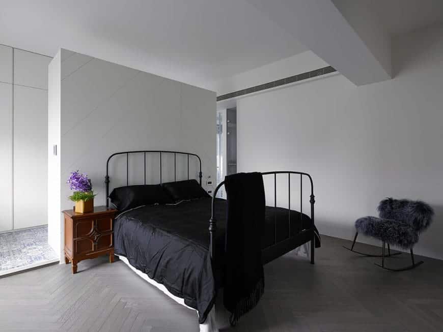 The black wrought iron bed frame matches with the black sheets that is complemented by the gray concrete flooring with a herring bone pattern to it contrasted by smooth white walls and ceiling.
