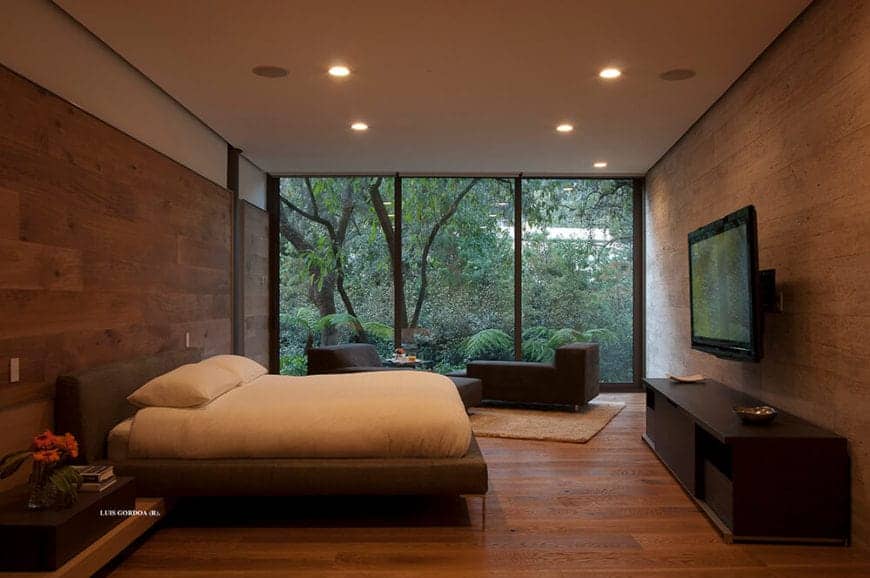 The white ceiling has recessed lights that cast down warm yellow lights onto the hardwood flooring that extends to the walls at the head and foot of the brown bed. This is given a nice background of lush greenery outside the glass wall.