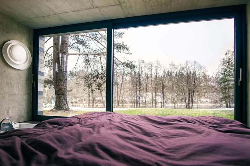 A whole wall of this primary bedroom is made of wide sliding glass doors that gives an unobstructed view of the surrounding landscape scenery of trees and grassy lawn. This serves as a nice background for the concrete gray walls and ceiling.