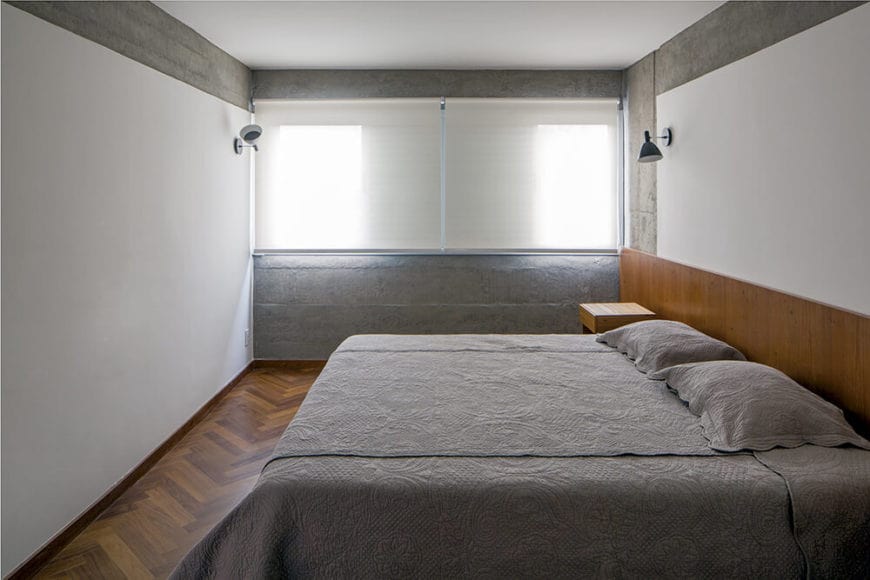The white walls and ceiling are accented with sections of gray concrete that matches with the gray sheets of the bed that has a wooden headboard extending to the bedside tables that matches with the hardwood flooring.
