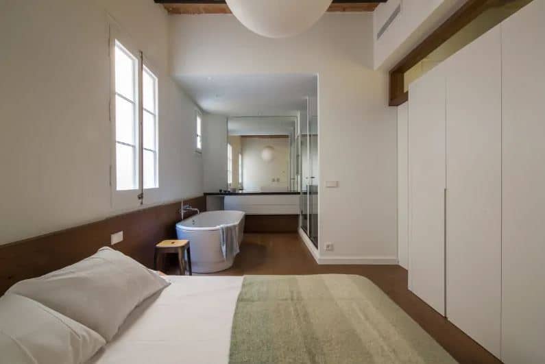 This is an industrial-style bedroom beside the bathroom on the same hardwood flooring that contrasts the white cushion with no bed frame. This hardwood flooring extends to a low wainscoting and matches with the wooden ceiling with exposed wooden beams.