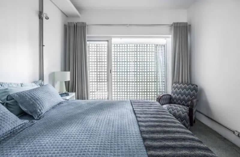 The light green textured and patterned sheets of the bed and its pillows contrast the white ceiling and walls accented with metal pipes that hold the wiring of the outlets and the spot light beside the bed.