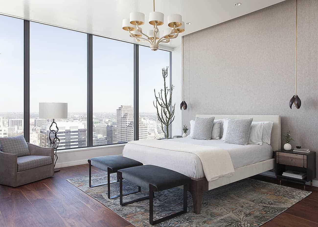 The dark hardwood flooring of this primary bedroom is brightened by the large glass walls on the right side of the bed that has a light gray headboard against a gray concrete wall accented with a pair of flower-like pendant lights.