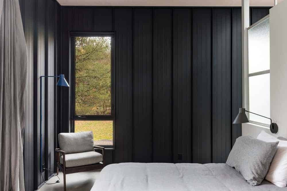 The light gray sheets of the bed matches with the gray concrete flooring and the cushions of the wooden armchair by the window that is inlaid into the industrial-style walls of dark gray material.