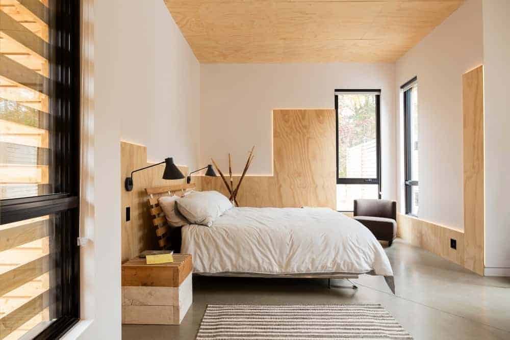 The ceiling of this primary bedroom is made of bare plywood that matches with the wooden accents of the white walls trimmed with back lighting to emphasize the lines. This is complemented by an industrial-style concrete flooring that goes well with the wooden bed flanked with repurposed wooden palette boxes.