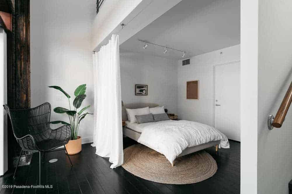 The industrial-style black hardwood flooring is topped with a rustic woven circular area rug underneath the gray bed that complements the white walls and the white curtain for privacy. This bed is topped with a row of spotlights mounted on the white ceiling.