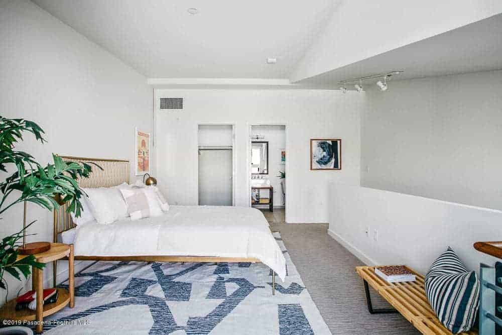 This primary bedroom has white walls and white ceiling that matches the white sheets of the bed that has an industrial-style brass frame that pairs well with the brass table lamps on the bedside tables flanking the bed over a gray carpeted flooring.