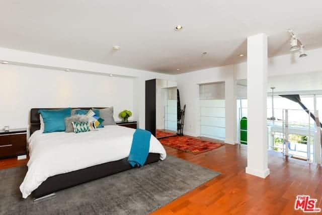 This is an industrial-style primary bedroom at a loft with white metal railings bordering one side with white support pillars that contrast the redwood flooring that is topped with an industrial-style gray area rug that complements the black leather bed with white sheets.