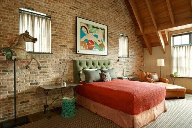 The green tufted headboard of the traditional bed is against a textured red brick wall adorned with a colorful painting flanked with square curtained windows. Beside the bed is a brown leather single sofa chair that matches with the wooden ceiling that has exposed wooden beams.