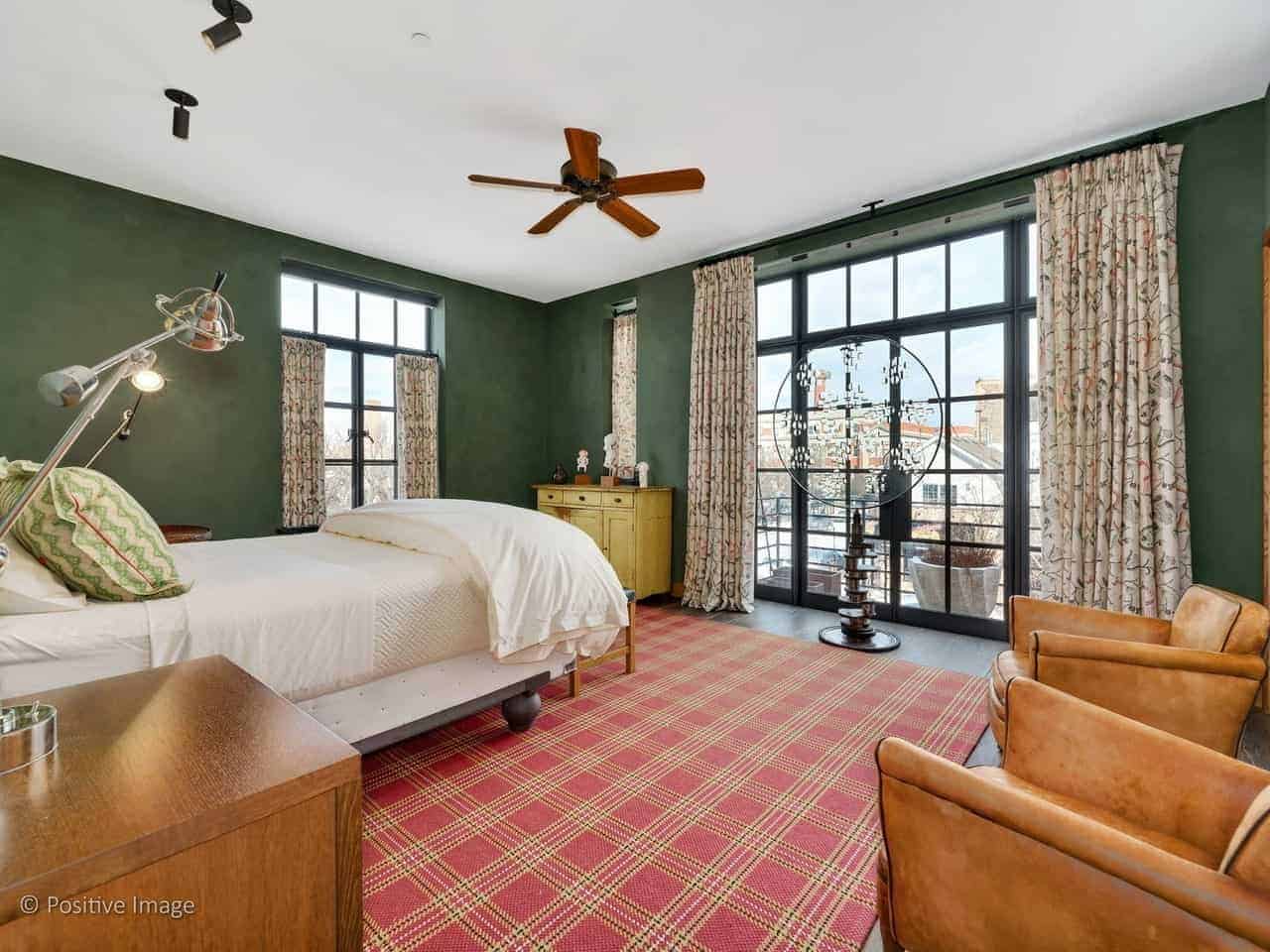 This industrial-style primary bedroom has dark green walls that are contrasted by the large French window and French glass doors. These are paired with floral curtains that pairs with the red plaid area rug to bring a dash of color to the bedroom.