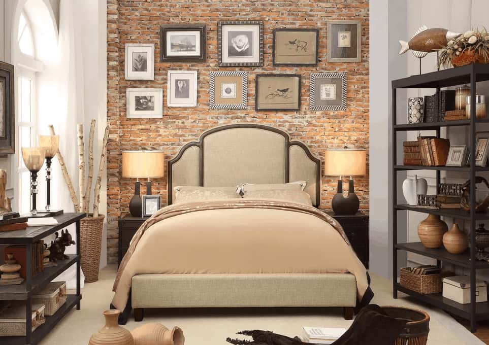 The beige sheets of the traditional bed goes well with the light gray cushioned headboard that stands out against the red brick wall behind it that is adorned with multiple framed photos and artworks. This is paired well with the shelves on either side of the bed that is filled with decors.