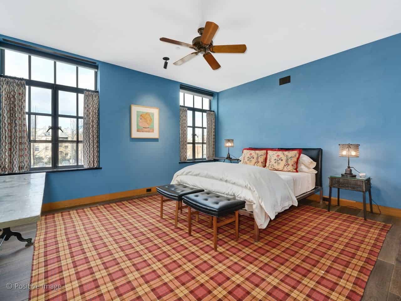 The hardwood flooring is covered with a red area rug that has a plaid pattern to it that provides a nice complex and colorful background for the black leather bed with stainless steel legs flanked with a couple of industrial-style bedside tables with matching table lamps.