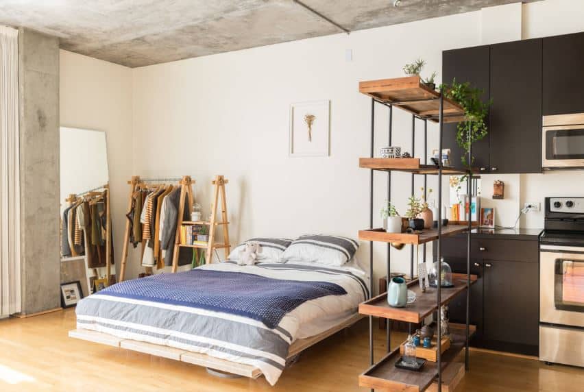 This bedroom is right beside the kitchen separated only by a wooden shelf with dark iron frames. This is complemented by a light hardwood flooring that blends with the platform bed and the clothes rack beside it.