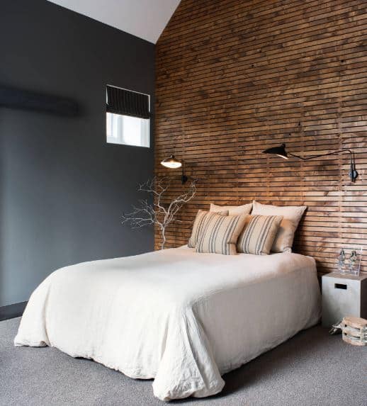 The gray carpeted flooring and gray wall on the side of the traditional bed is complemented by the wall at the head of the bed. It has an industrial-style wooden panel finish that supports a couple of wall-mounted lamps.