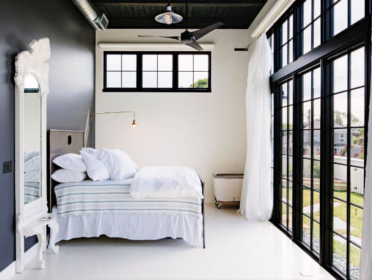 The brightness of this bedroom is due to the natural lights coming in from the French glass doors that brighten the white flooring and wall adjacent to the black headboard of the traditional bed topped with a ceiling with exposed beams, vents and tubes.