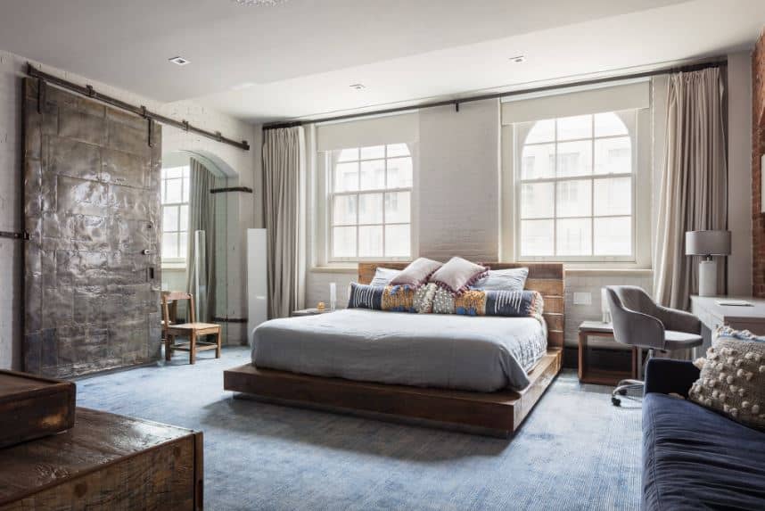 The blue distressed carpeting of the floor is a nice complement to the wooden platform with a plank finish to its wooden headboard that stands out against the white walls brightened by the natural lights of the arched windows.