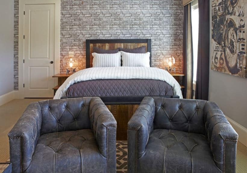 The couple of worn out gray tufted armchairs at the foot of the bed goes well with the industrial-style aesthetic of the bedroom that has gray brick walls behind the wooden headboard of the bed that looks like it came from a barrel.