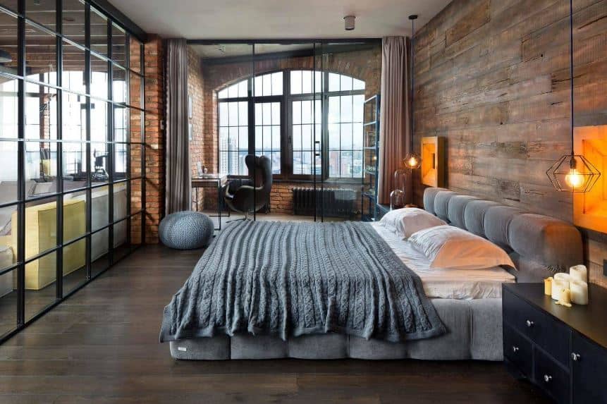 This industrial-style primary bedroom is separated from the rest of the house with glass walls that go well with the dark hardwood flooring. On the far wall is a small office with a large arched window and red brick walls that match the wooden wall of the gray cushioned headboard.