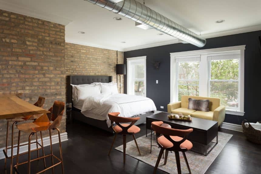 The black hardwood flooring is matched with the black wall that supports the white framed windows topped with a stainless steel exposed vent of the white ceiling contrasted by brick walls near the dark gray cushioned bed.