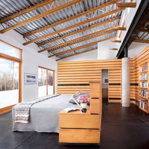 The gray bed sits in the middle of the black industrial flooring contrasted by the light wood headboard and bedside drawer of the bed as well as the walls surrounding the bed. This is topped with a factory-like ceiling of metal beams and tin roofing.