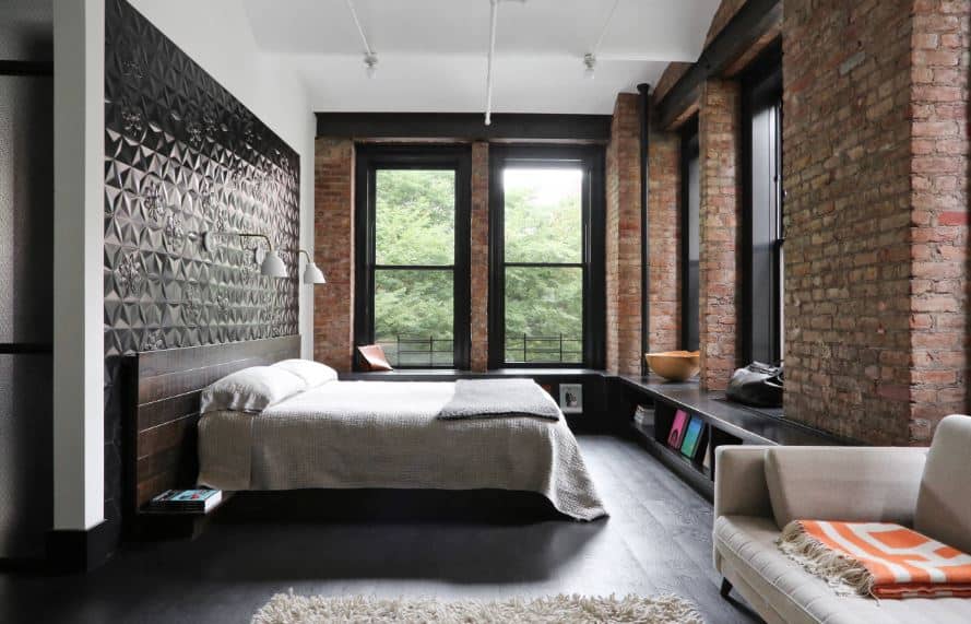This industrial-style primary bedroom is surrounded with tall windows that are supported by brick pillars framed with black wooden shelves at the bottom that matches with the black wall panel behind the wooden headboard.