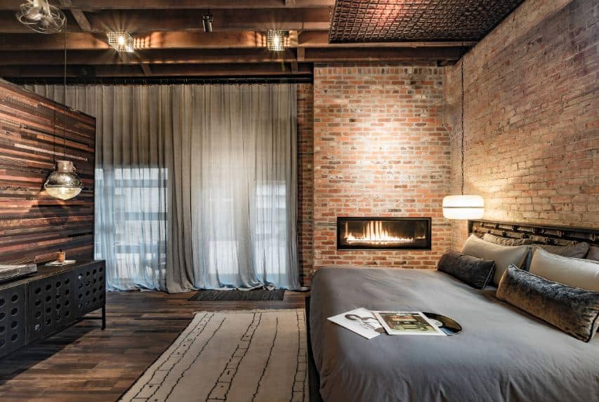 The comfortable gray bed is lit with a warmth from the round pendant light augmented by the fireplace on the side that is inlaid in a red brick wall topped with a ceiling filled with exposed wooden beams matching the hardwood flooring.