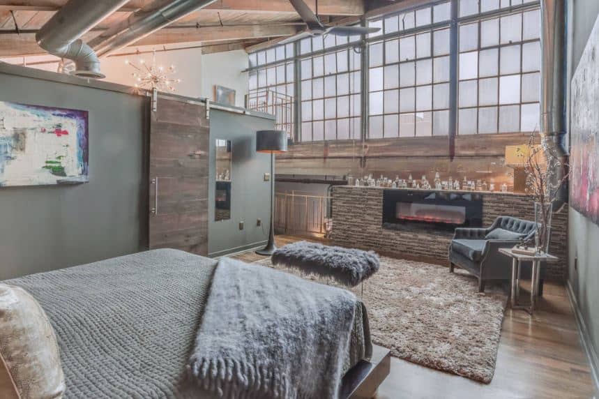This bedroom looks like its part of a renovated factory that has a wooden shed ceiling with exposed beams, pipes and vents paired with large windows filled with frosted glass and a modern fireplace by the foot of the bed that has a sitting area.
