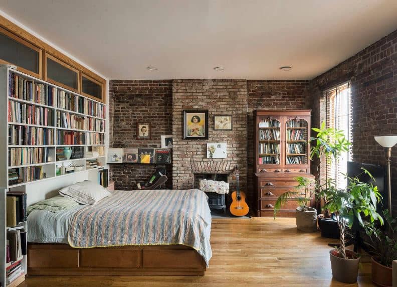 This cozy and homey industrial-style bedroom has a headboard with built-in bookshelves adjacent to a wall of red bricks that houses a fireplace and an antique bookshelf and dresser that matches with the wooden frame of the bed.