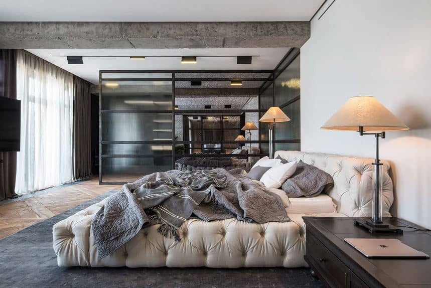 The cushion is placed on a platform bed frame that is upholstered with white leather and given a tufted finish that extends to the headboard. This stands out against the gray area rug that matches with the exposed concrete beam of the ceiling.