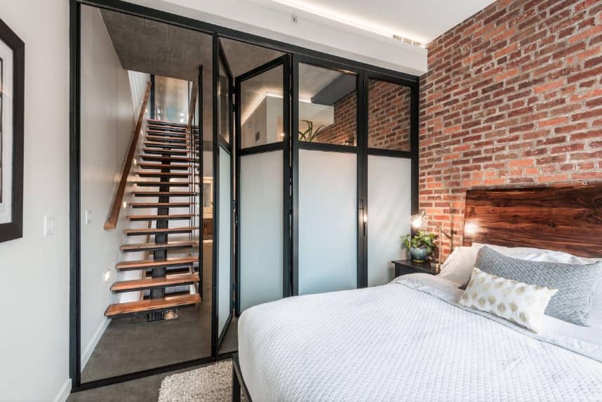 This small industrial style bedroom has a wall of frosted glass that folds to open to the wooden staircase. Adjacent to this is the red brick wall behind the wooden headboard of the bed over a concrete floor with a beige area rug.
