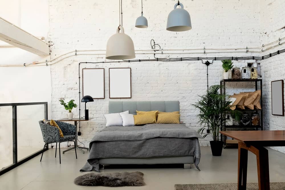 The exposed wiring of this industrial-style bedroom stands out against the white stone textured wall behind the gray cushioned headboard that matches with the gray sheets that stand out against the light gray flooring.