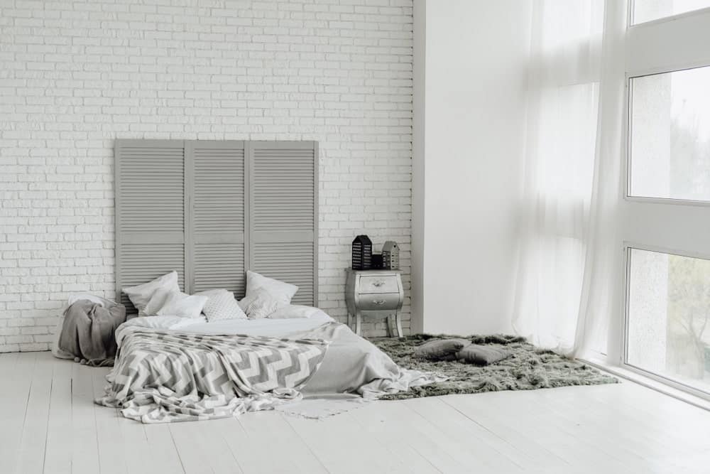 The sheer curtained glass windows bring in an abundance of natural lighting that brightens up the white textured wall with a brick wall pattern. This is complemented by wooden divider that is used as a tall headboard for the platform bed on the light gray flooring.
