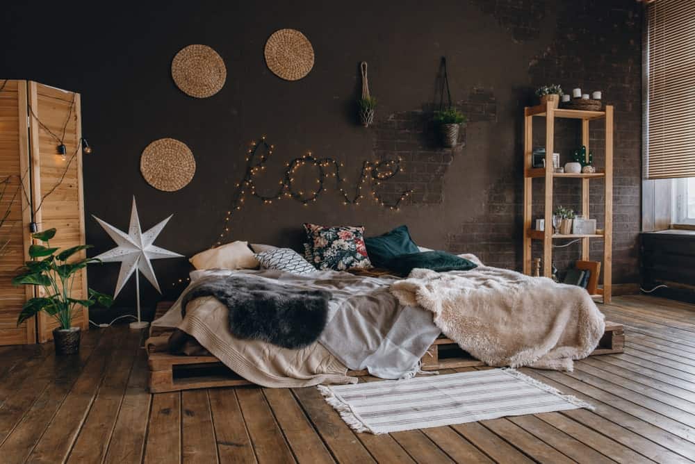 This industrial-style primary bedroom has a romantic vibe to it with a platform bed frame made of pallet crates that blend with the distressed hardwood flooring and complements the wooden structures of the room that stand out against the dark gray wall.