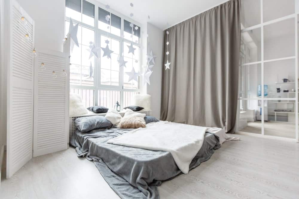 The high white ceiling of this industrial-style bedroom seems higher with a bed right down on the light hardwood flooring. This is augmented by a tall window at the head of the bed with a wooden headboard that also serves as a sitting area by the window.