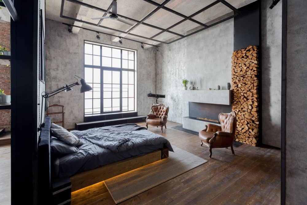 This industrial-style bedroom has a warm fireplace embedded into a gray concrete wall with a firewood storage beside it. The wooden traditional bed across from it has a back lighting underneath that gives the hardwood flooring and woven area rug an ethereal yellow glow.