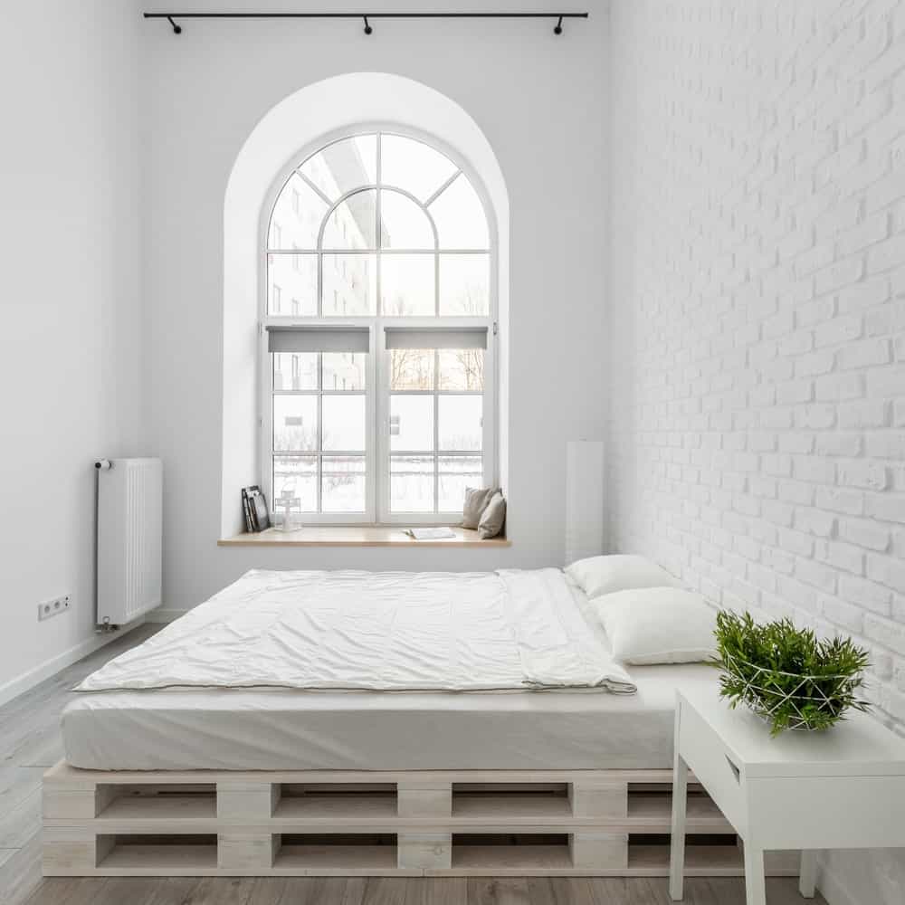 The brightness of this industrial-style bedroom comes from the tall arched window that brings in an abundance of natural lighting that makes the white walls and bed glow. This bed is on a stack of pallets forming a platform for the bed cushion.