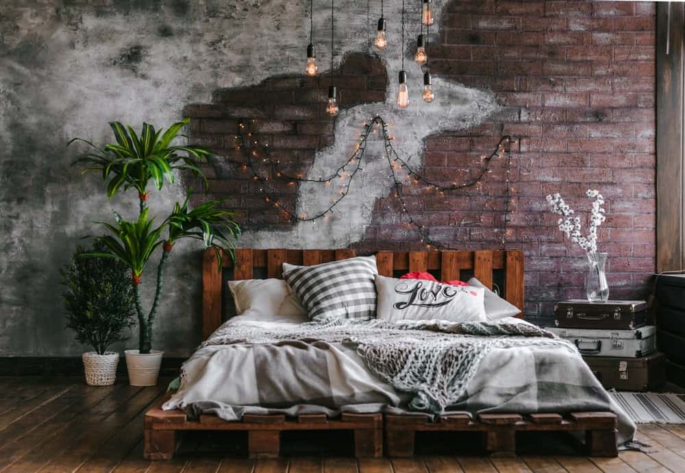 This industrial-style bedroom has a customized platform bed frame made of wooden pallet crates that also make up the wooden headboard with a redwood hue. This complements the red brick wall with a design that fades into a concrete wall.