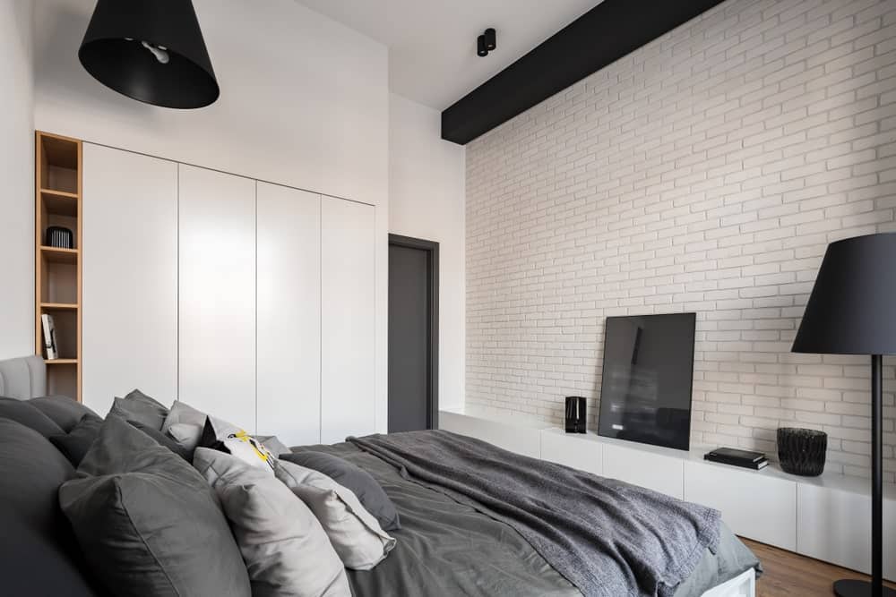 The dark gray sheets and pillows of the traditional bed stands out against this predominantly white primary bedroom that has white brick walls, white cabinets embedded into the white walls and a white ceiling that is contrasted by the black pendant light and beam.