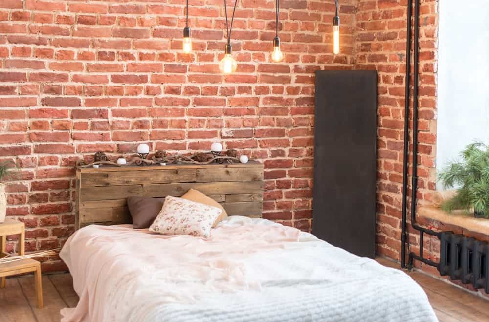The rustic wooden headboard of the platform-style bed matches the hardwood flooring and its plank finish. This is surrounded by red brick walls that are accented with black pipes of the heater and a variety of pendant lights.