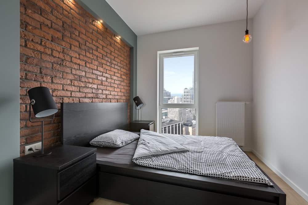 The simplicity of this industrial-style bedroom brings focus to its red brick wall that complements the black wooden headboard of the platform-style black wooden bed. This matches with the black wooden bedside drawers and its table lamps.