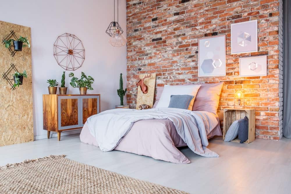 This charming industrial-style bedroom has a chic quality to its aesthetic of pink bed sheets, red brick wall and a palette crate bedside table that also acts as pillow storage. The red brick wall is adorned with white artworks that match the flooring.
