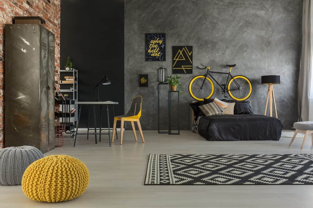 The industrial-style gray concrete wall provides a nice complement to the black sheets of the low bed with a wooden headboard that also serves as the stand for the bicycle. This acts as an accent to the gray wall along with framed artworks and a standing lamp.