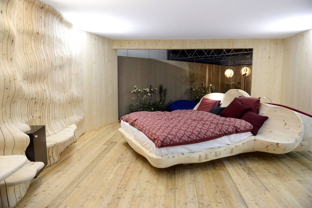 This industrial-style bedroom has light hardwood flooring that blends with the unique customized bed frame that has a fluid shape. This aesthetic is matched by the wall of the same material and hue that seem to flow and ripple to create shelves.