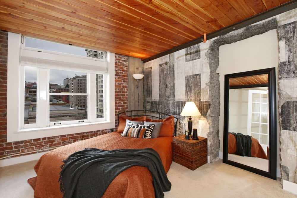 The four surfaces of this industrial-style bedroom has a different hue and material. The flooring is beige, the two adjacent walls flanking the bed is also varied with the left side made of brick and the right made of concrete and finally the ceiling is made of wood.