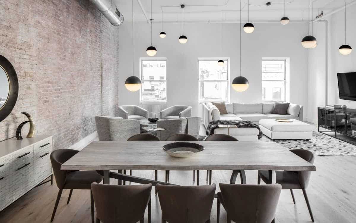 The wooden dining table of this industrial-style dining room matches with the hardwood flooring contrasted by the dark brown cushioned chairs. The tall red brick wall complements this and the exposed vent of the white ceiling.