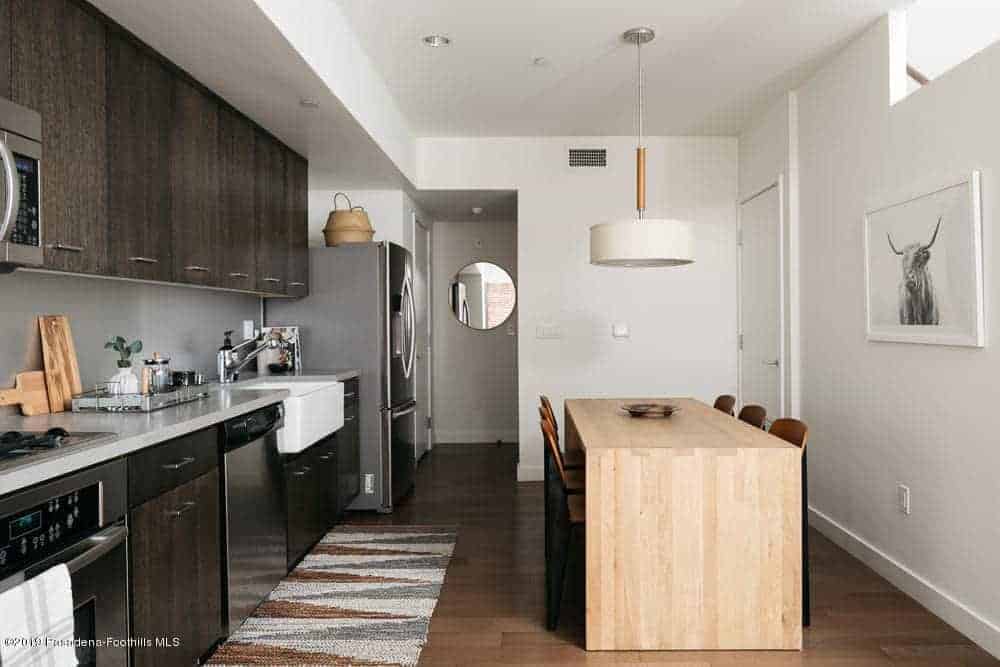 The simple waterfall wooden dining table of this informal dining area stands out against the dark hardwood flooring but complements the white ceiling that hangs a circular white pendant light over the table and its wooden chairs.
