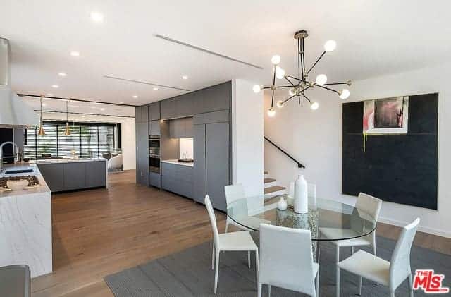 The circular glass-top dining table over the gray area rug is paired with modern white dining chairs that match the white ceiling that supports an industrial-style chandelier casting yellow warm lights.