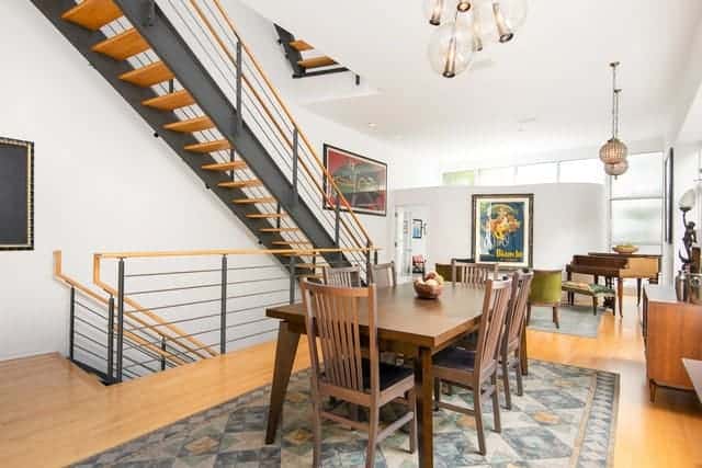 This dining area has a background of the metal railings and metal beams of the staircase that stands out against the light hardwood flooring topped with a distressed patterned area rug underneath the wooden dining set.